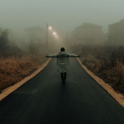 A person stands with outstretched arms on a foggy road in a misty neighborhood, creating a moody atmosphere.